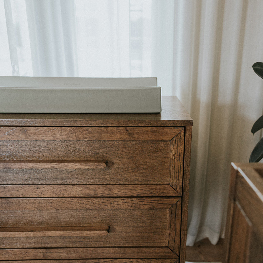 A close-up of a dark brown wooden dresser with a light gray changing pad on top, set against a background of white curtains and a green houseplant.